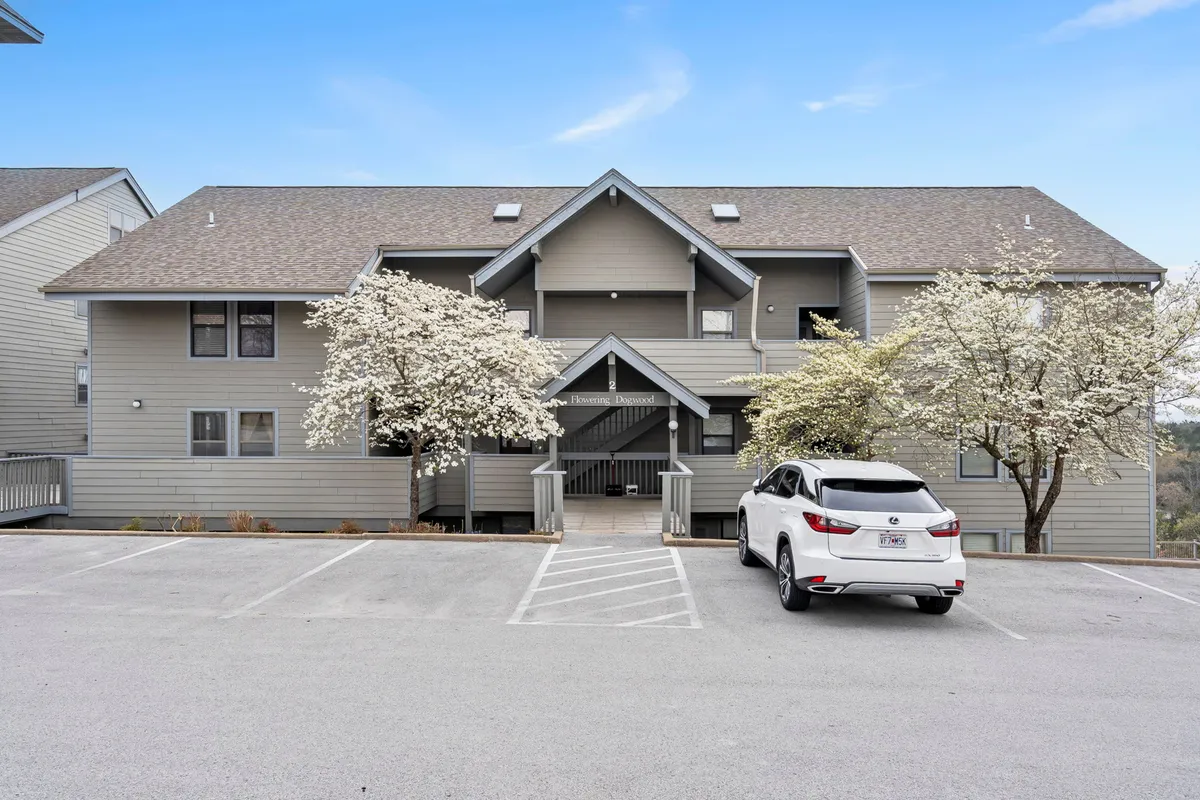 This exterior image features flowering trees, an inviting entrance porch, and a vehicle in the parking lot, presenting a welcoming view of the property. The presence of greenery and accessible walkways highlights ease of access and a peaceful, residential environment.