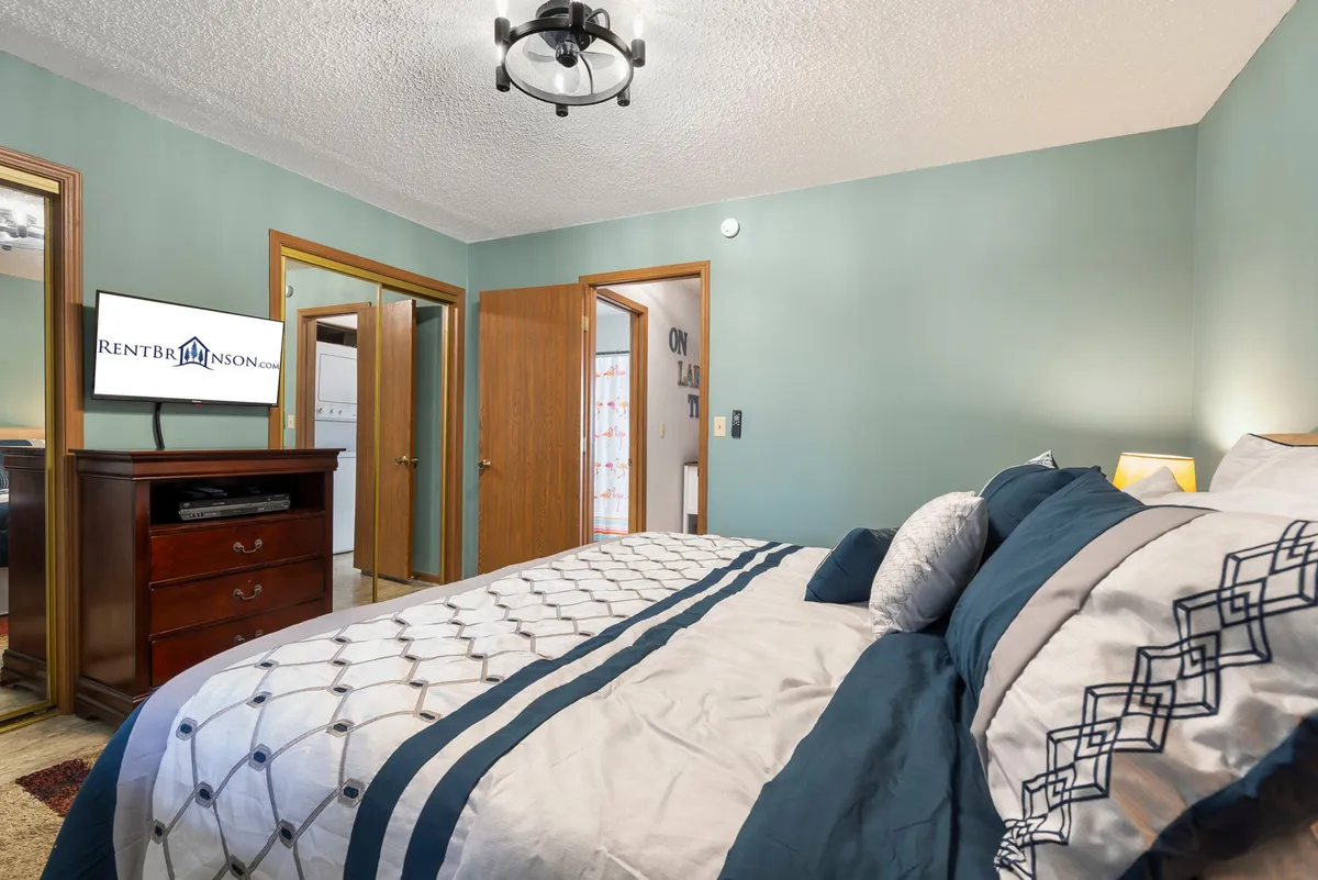 A cozy, well-appointed bedroom featuring a king bed with decorative bedding, a wooden dresser, and a TV mounted on the wall. The room is spacious, with open doors leading to other areas, and soft, inviting lighting. The overall atmosphere is restful and comfortable, ideal for unwinding after a busy day.