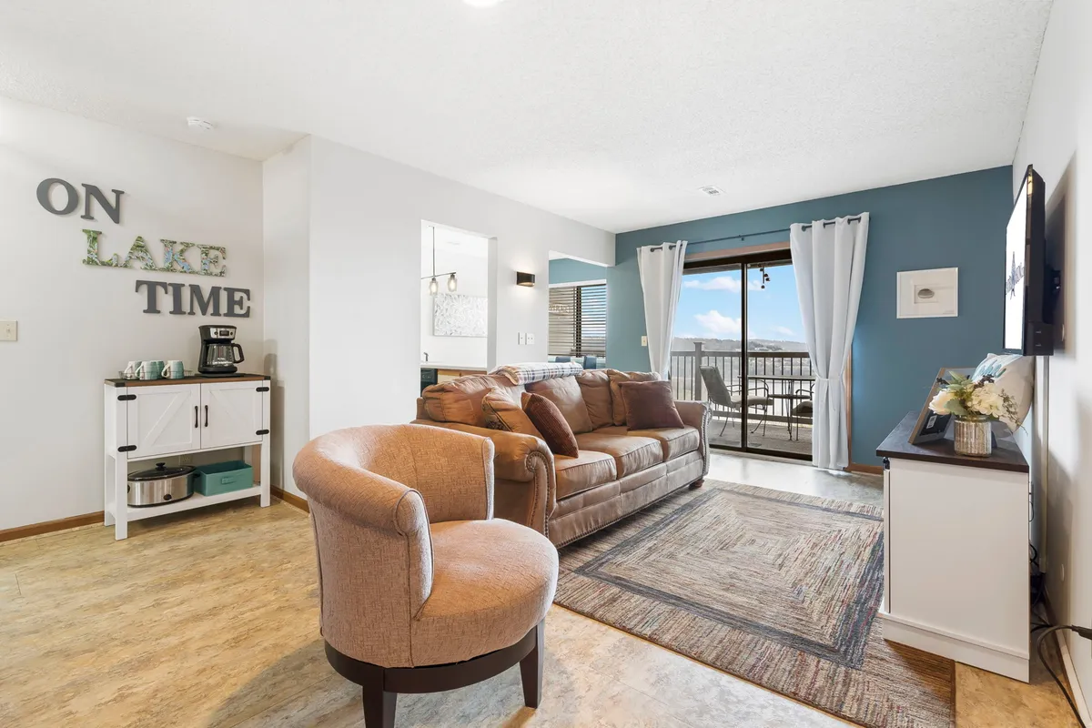 A spacious, sunlit living room featuring a comfortable sofa and accent chair oriented toward a large window that offers expansive views of Table Rock Lake. Decorative wall art adds a touch of personality, and the setup invites family relaxation with the possibility of gathering for conversation, coffee, or TV time.