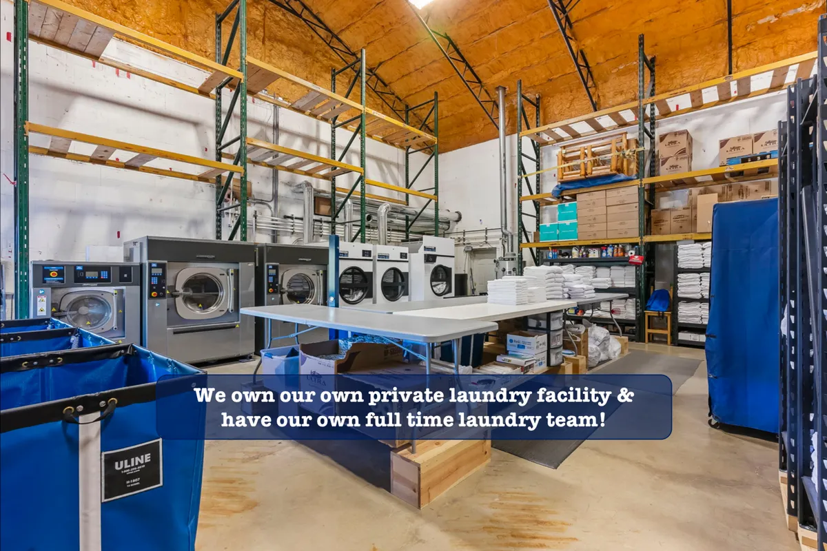 An interior shot of a professional laundry room with industrial washing machines, folding tables, and organized shelves. The scene emphasizes the high cleaning standards and attention to detail provided by the property's management.
