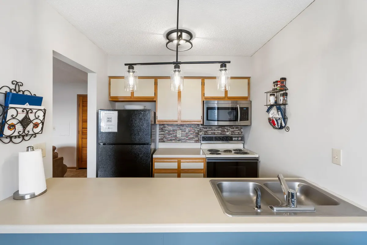 This image features a modern kitchen with a prominent kitchen island and sink, a black refrigerator, stainless steel appliances, and stylish hanging light fixtures. A decorative wall shelf adds personality, while the layout suggests a functional and inviting cooking space ideal for meal preparation and entertaining.