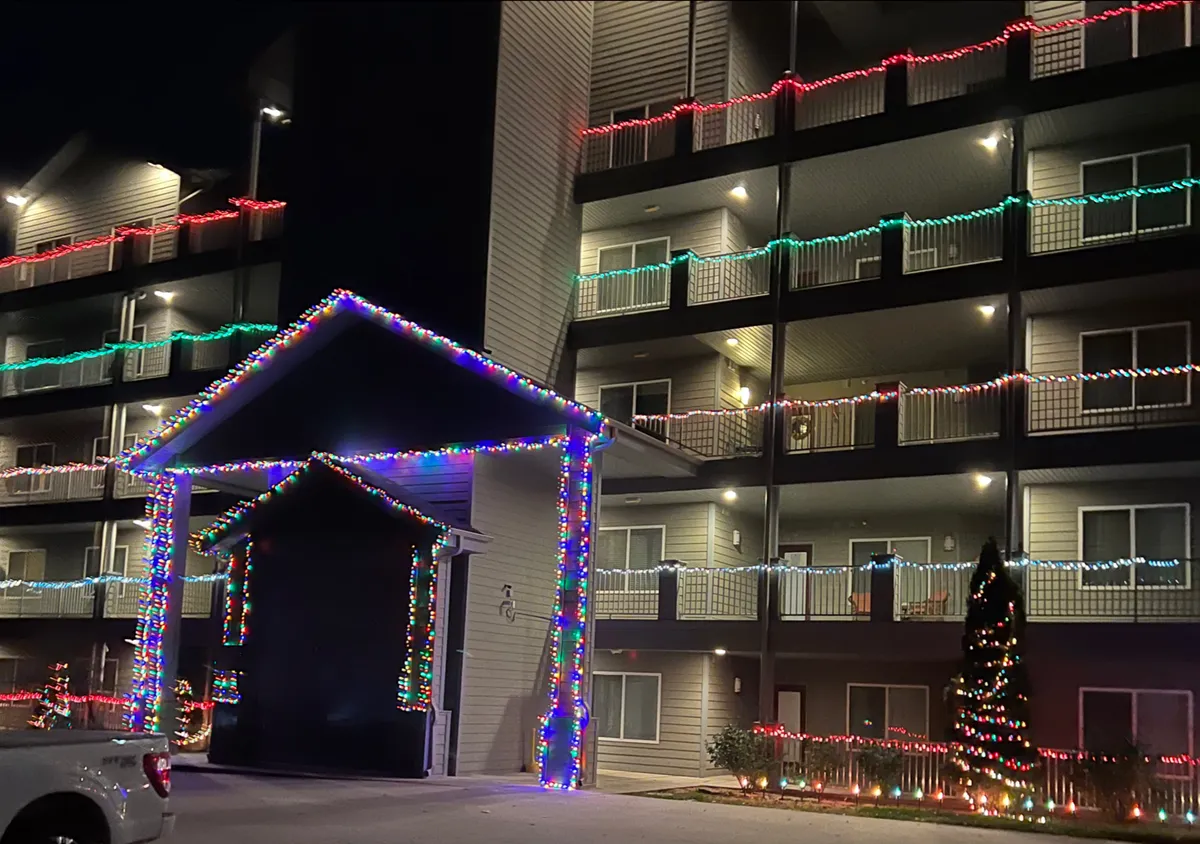 An exterior view at dusk shows the property decorated with festive lights, balconies, and a holiday tree, conveying a joyful and community-focused atmosphere. The soft lighting and inviting common areas suggest relaxing evenings and opportunities for social enjoyment.