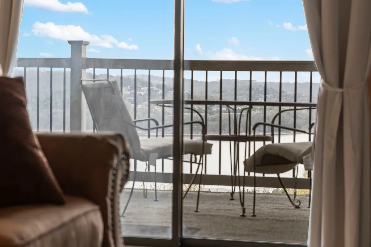 A tranquil interior scene featuring a table and chairs set near a sliding door that opens to a private balcony. Sheer curtains frame the access point to the deck, while a sofa nearby suggests easy movement between indoor relaxation and outdoor enjoyment. The balcony overlooks natural surroundings, reinforcing the connection to the lake and fresh air.
